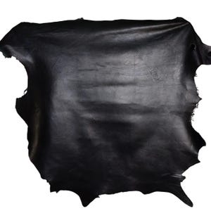 May include: A large, irregularly shaped piece of black leather. The leather has a smooth, slightly shiny surface with natural markings and variations in texture. The edges are uneven, showing the raw cut of the material. The overall appearance suggests a high-quality, natural material.