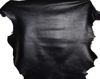 May include: A large, irregularly shaped piece of black leather. The leather has a smooth, slightly shiny surface with natural markings and variations in texture. The edges are uneven, showing the raw cut of the material. The overall appearance suggests a high-quality, natural material.