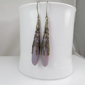 May include: A pair of dangle earrings with a light purple teardrop-shaped glass bead encased in a gold filigree design.