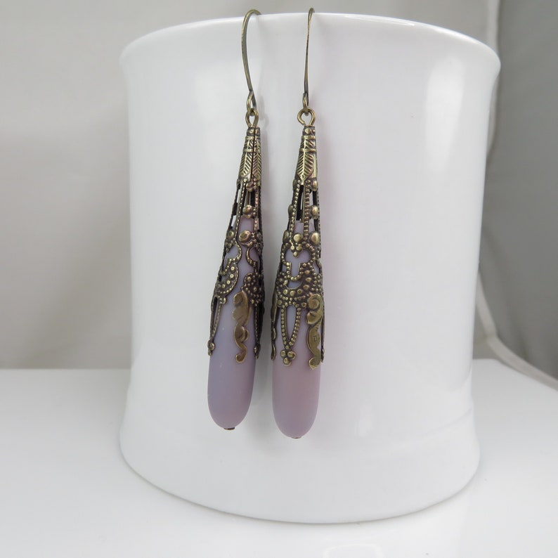 May include: A pair of dangle earrings with a light purple teardrop-shaped glass bead encased in a gold filigree design.