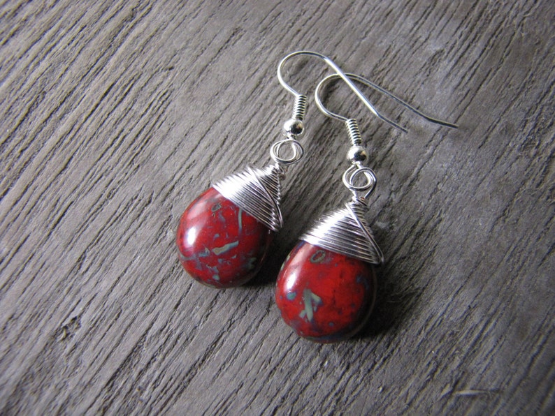 May include: A pair of teardrop-shaped earrings. Each earring features a red stone with green and gray accents, wrapped with silver wire. The earrings hang from silver-colored ear wires, showcasing a handcrafted jewelry design.