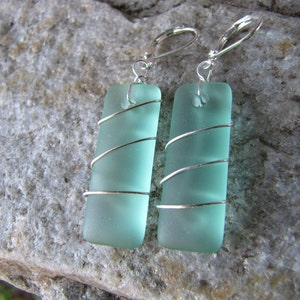 May include: A pair of rectangular, seafoam green earrings. Each earring features a rectangular piece of frosted glass wrapped with silver wire. The earrings have silver lever-back closures. The earrings are displayed on a gray stone surface.