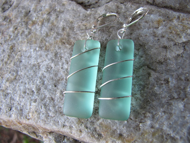 May include: A pair of rectangular, seafoam green earrings. Each earring features a rectangular piece of frosted glass wrapped with silver wire. The earrings have silver lever-back closures. The earrings are displayed on a gray stone surface.