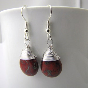 May include: A pair of handcrafted earrings featuring teardrop-shaped red and green gemstones. The stones are wrapped with silver wire, and the earrings have silver-colored ear wires. The earrings are displayed against a white background.