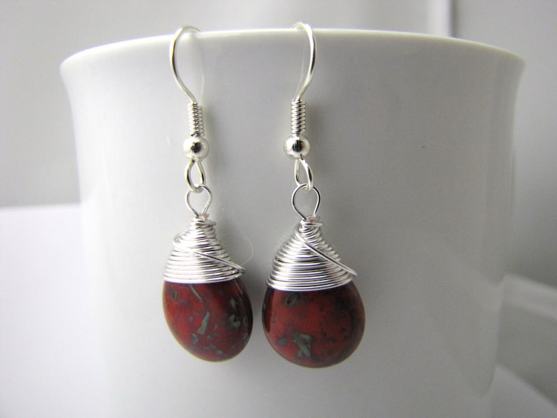 May include: A pair of handcrafted earrings featuring teardrop-shaped red and green gemstones. The stones are wrapped with silver wire, and the earrings have silver-colored ear wires. The earrings are displayed against a white background.