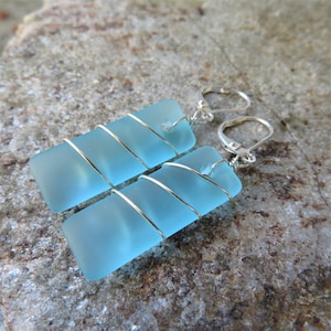 May include: A pair of light blue rectangular earrings. Each earring features a rectangular piece of sea glass wrapped with silver-colored wire. The earrings have silver-colored lever-back closures. The earrings are displayed on a textured, gray surface.