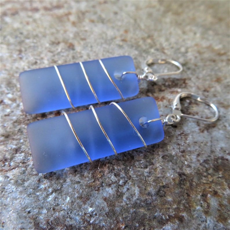 May include: A pair of rectangular blue earrings with a frosted finish. Each earring is wrapped with silver-colored wire in a spiral pattern. The earrings have silver-colored lever-back closures. The jewelry is displayed on a textured, neutral-toned surface.