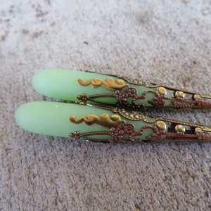May include: A pair of green glass teardrop-shaped beads with ornate gold metal caps. The caps have a filigree design with swirls and dots.