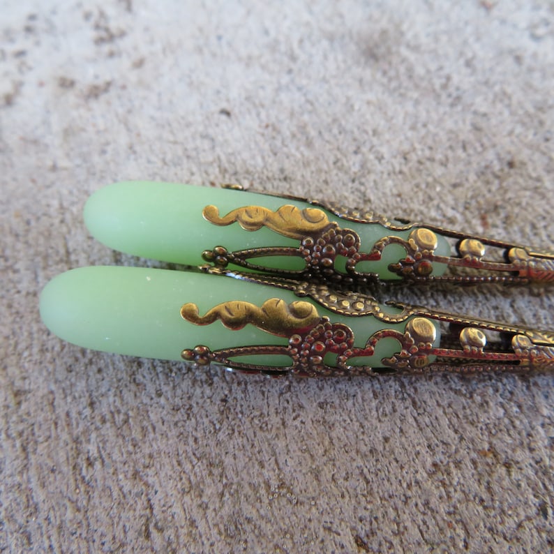 May include: A pair of green glass teardrop-shaped beads with ornate gold metal caps. The caps have a filigree design with swirls and dots.