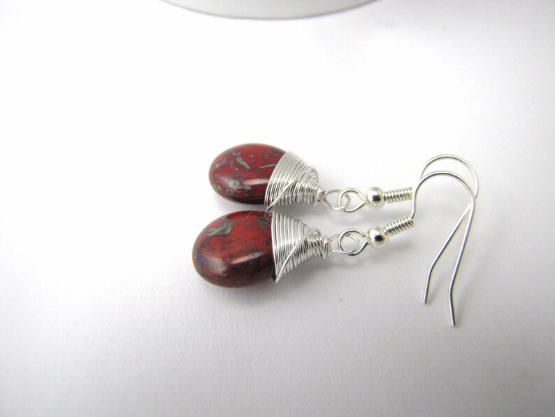 May include: A pair of dangle earrings featuring oval-shaped, red and green speckled stones. The stones are wrapped with silver wire and attached to silver-colored fish hook ear wires. The earrings are displayed on a white surface.