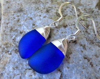May include: Cobalt blue glass earrings with silver wire wrapped around the top of each teardrop-shaped piece. The earrings hang from silver hooks.