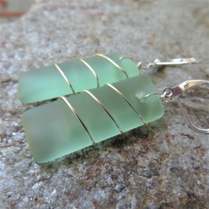 May include: Sea glass earrings with a rectangular shape and a pale green hue. The sea glass is wrapped with gold-colored wire and attached to silver-colored leverback earring hooks. The earrings are displayed on a textured, gray surface.