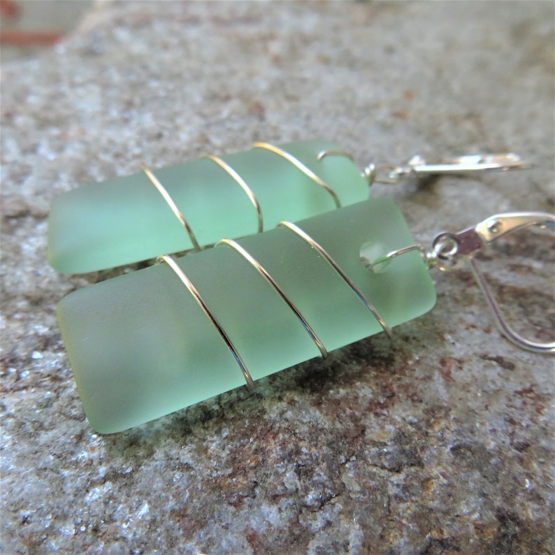 May include: Sea glass earrings with a rectangular shape and a pale green hue. The sea glass is wrapped with gold-colored wire and attached to silver-colored leverback earring hooks. The earrings are displayed on a textured, gray surface.
