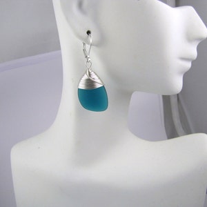 May include: Teal blue glass teardrop shaped earrings with silver wire wrapping. The earrings are on a white mannequin head.