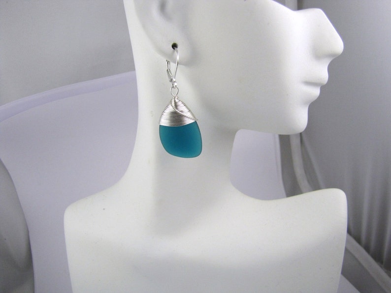 May include: Teal blue glass teardrop shaped earrings with silver wire wrapping. The earrings are on a white mannequin head.