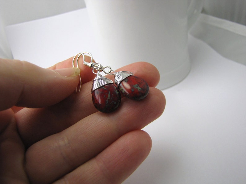 May include: A pair of teardrop-shaped earrings with a red and green speckled pattern. The earrings are wire-wrapped with silver-colored metal and have silver-colored ear hooks. The earrings are displayed against a white background.