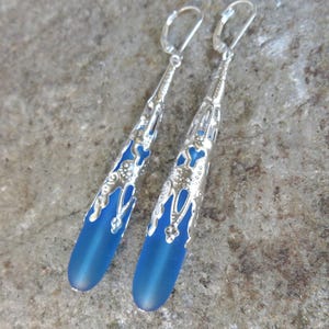 May include: A pair of long, silver dangle earrings with blue glass teardrop-shaped beads. The beads have a frosted finish and are set in a silver filigree design.