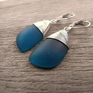 May include: Teal blue glass earrings with silver wire wrapped around the top of each earring. The earrings are hanging from silver hooks.