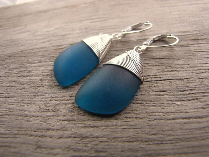 May include: Teal blue glass earrings with silver wire wrapped around the top of each earring. The earrings are hanging from silver hooks.