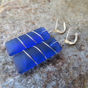 May include: A pair of rectangular blue earrings. Each earring features a piece of blue glass wrapped with silver wire. The earrings have silver lever-back closures. The earrings are displayed on a textured, gray surface.