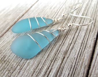 May include: A pair of drop earrings featuring smooth, frosted light blue sea glass pieces. Each piece of glass is wrapped in thin, polished silver-colored wire and attached to a simple silver-colored metal hook for hanging from the ear.