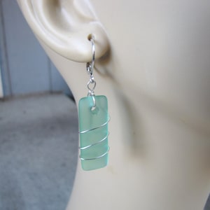 May include: A pair of rectangular sea glass earrings. Each earring features a light green, rectangular piece of sea glass wrapped with silver-colored wire. The earrings have a lever-back closure.