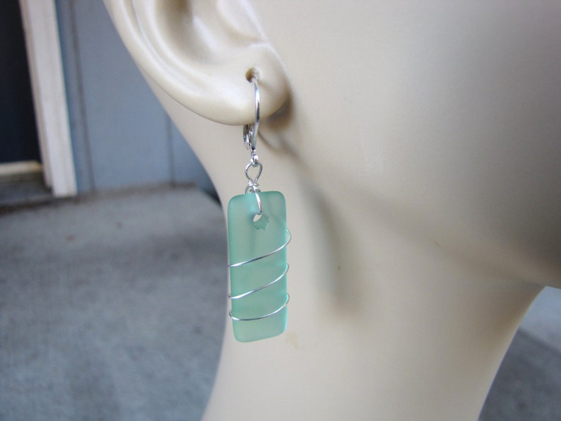 May include: A pair of rectangular sea glass earrings. Each earring features a light green, rectangular piece of sea glass wrapped with silver-colored wire. The earrings have a lever-back closure.