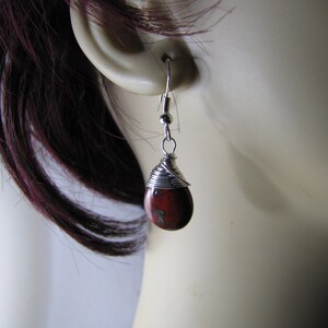 May include: A single teardrop-shaped earring with a red and brown stone, wrapped in silver wire. The earring hangs from a silver hook. The earring is shown against a blurred background.