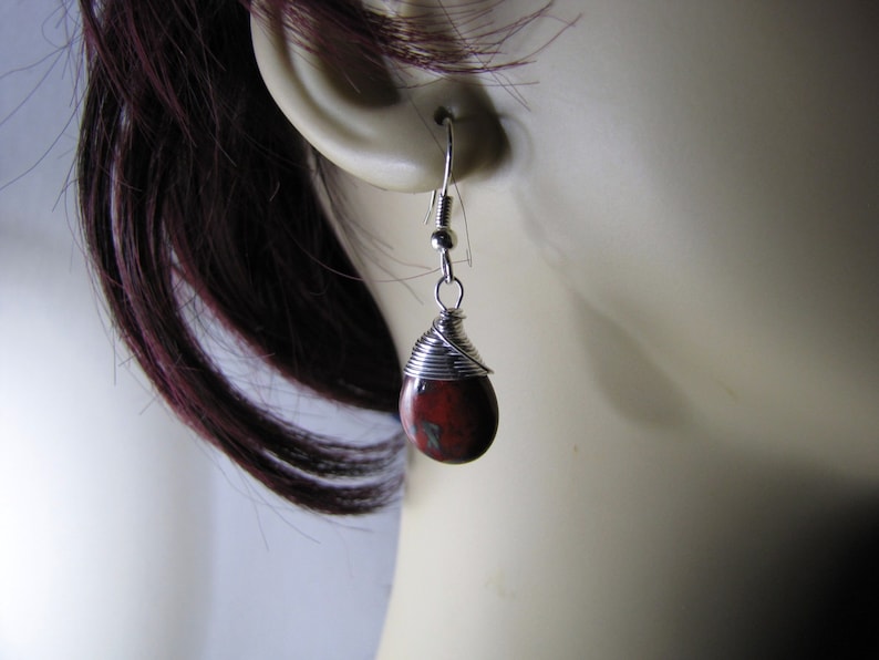May include: A single teardrop-shaped earring with a red and brown stone, wrapped in silver wire. The earring hangs from a silver hook. The earring is shown against a blurred background.