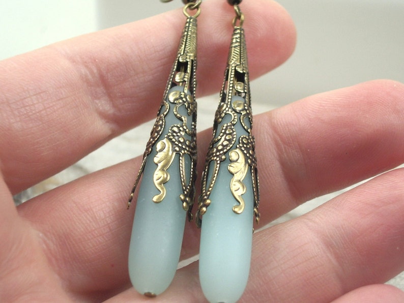 May include: A pair of dangle earrings with a light blue glass teardrop shape and a gold filigree design.