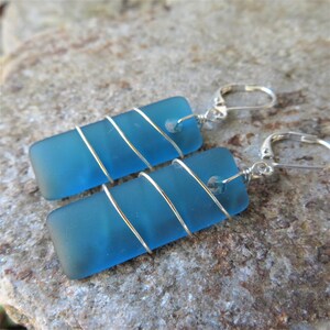 May include: A pair of rectangular blue earrings. Each earring features a piece of sea glass wrapped with silver wire. The earrings have silver lever-back closures. The sea glass has a frosted appearance.