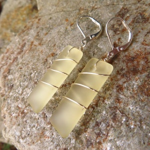 May include: A pair of rectangular earrings with a pale yellow, frosted glass appearance. Each earring is wrapped with silver wire and has a silver lever-back closure. The earrings are displayed on a textured, gray-brown stone surface.