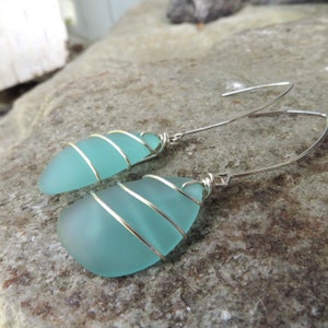 May include: A pair of silver wire wrapped earrings with light blue sea glass. The earrings are dangle style and have a simple design.