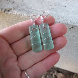 May include: A pair of rectangular, seafoam green earrings. Each earring is wrapped with silver wire and has a silver lever-back closure. The earrings are rectangular and have a frosted appearance.