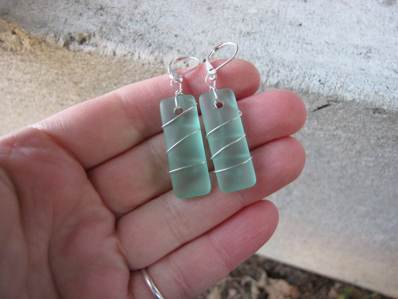 May include: A pair of rectangular, seafoam green earrings. Each earring is wrapped with silver wire and has a silver lever-back closure. The earrings are rectangular and have a frosted appearance.