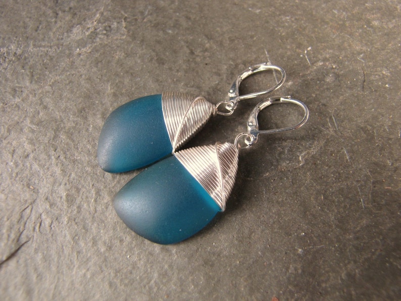 May include: Teal blue glass earrings with silver wire wrapped around the edges. The earrings have silver hooks.