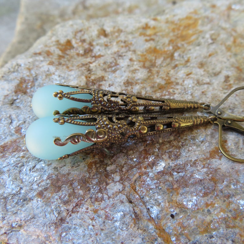 May include: A pair of long, dangling earrings with a delicate, ornate design. The earrings feature a light blue, frosted glass bead at the bottom of each earring. The metal is a brassy gold color.