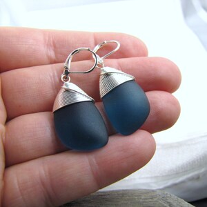 May include: A pair of dangle earrings featuring dark blue sea glass wrapped in silver wire.