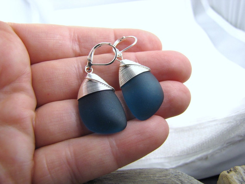 May include: A pair of dangle earrings featuring dark blue sea glass wrapped in silver wire.