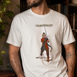 Peut inclure: Un t-shirt blanc à manches courtes orné d'une illustration graphique représentant une personne en tenue médiévale tenant une longue épée. Au-dessus de la figure se trouve une bannière avec le texte "Posta Frontale" et un petit chat blanc.