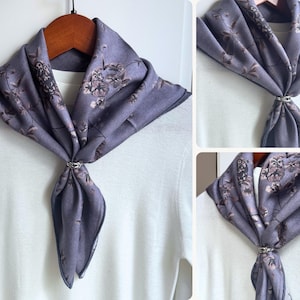May include: A lavender scarf with a floral pattern, secured with a silver-colored ring. The scarf is draped over a white top, creating a stylish accessory. The scarf is made of a soft material.