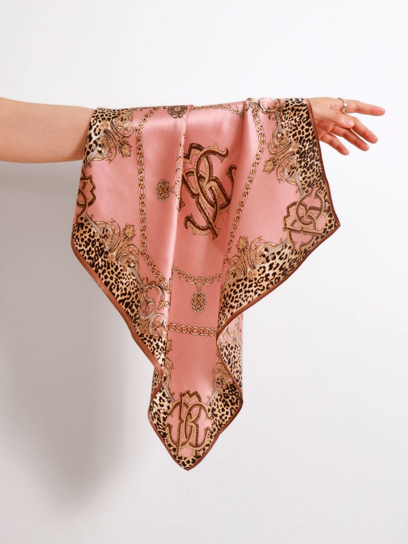 May include: A pink silk scarf with a brown leopard print border and gold chain and scrollwork details. The scarf features a large monogram in the center. The scarf is held up by a person's hand.