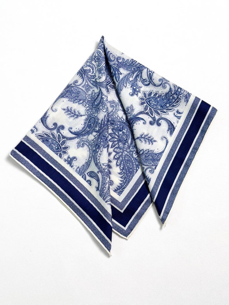 May include: White and blue paisley patterned handkerchief. The handkerchief features a detailed floral design in shades of blue against a white background. The edges are trimmed with a navy blue stripe and a thin white stripe.
