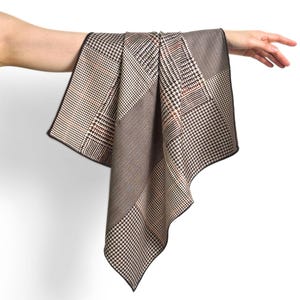 Puede incluir: Un pañuelo cuadrado con un diseño de patchwork de patrones marrones, negros y crema, incluyendo pata de gallo y espiga. La tela tiene un borde acabado en marrón oscuro y está drapeada para mostrar claramente las texturas textiles geométricas.
