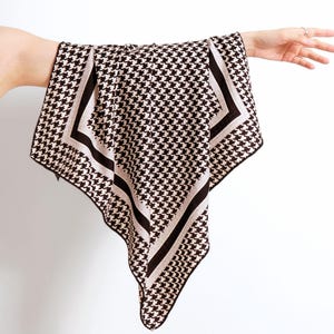 Puede incluir: Un pañuelo con estampado de pata de gallo marrón y blanco. El pañuelo tiene forma cuadrada con un borde contrastante negro y beige. El pañuelo se sostiene sobre un fondo blanco.