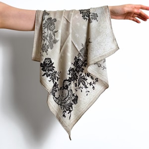 May include: A beige square scarf with black floral lace patterns. The scarf is held up by a hand, showcasing the intricate design and delicate fabric. The edges feature a detailed border.