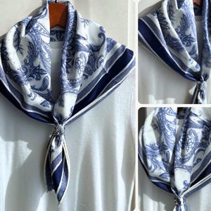 May include: A white and blue paisley patterned scarf with a dark blue border. The scarf is knotted at the front and draped over a white top. The scarf is made of a soft, lightweight fabric.