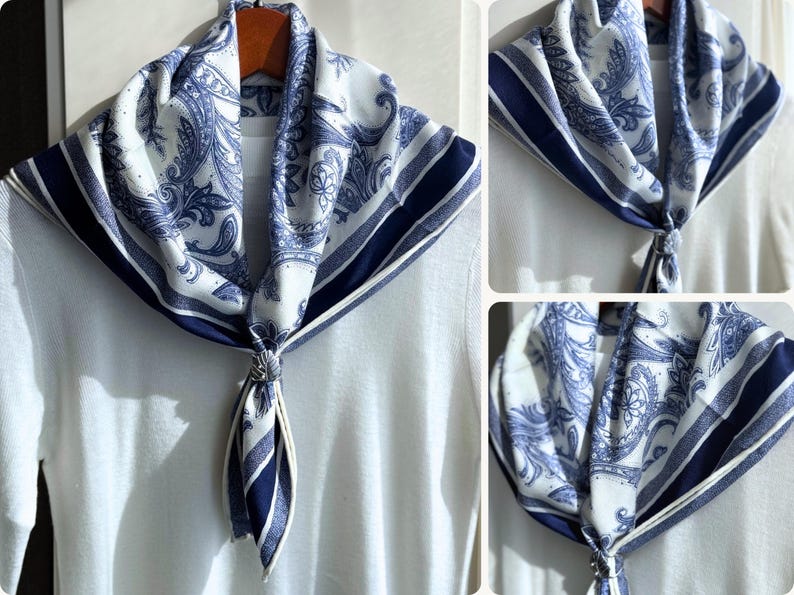 May include: A white and blue paisley patterned scarf with a dark blue border. The scarf is knotted at the front and draped over a white top. The scarf is made of a soft, lightweight fabric.