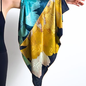 May include: A silk scarf with a floral pattern in teal, yellow, and navy blue. The scarf is held up, displaying its design. The floral pattern is detailed and covers the entire surface. The scarf's edges are finished with a navy border, adding a touch of elegance.