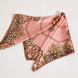 May include: A pink satin scarf with a gold chain and baroque design. The scarf has a leopard print border and a brown trim. The scarf is folded and displayed on a white surface.